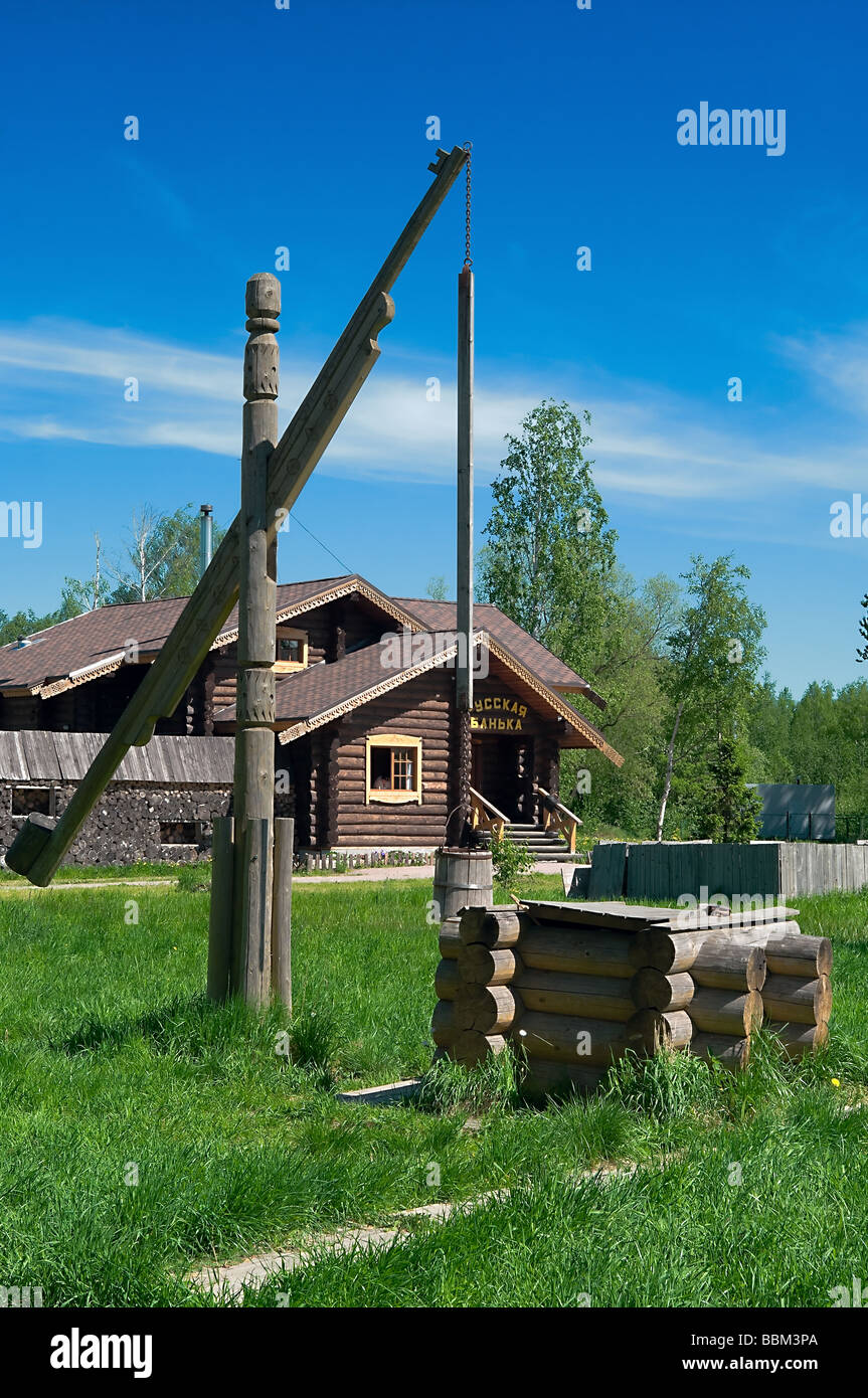 Wooden draw well in the rural area with shadoof. Green grass around it. Wooden house in background. Russian village. Russia Stock Photo