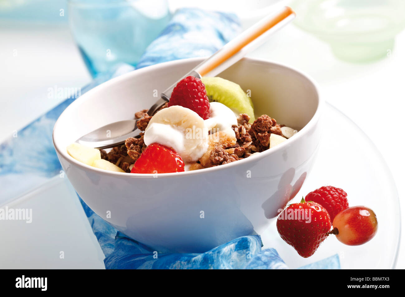 Muesli in a small porcelain bowl with yoghurt, fruit, caramel brittle, raspberries, strawberries, grapes, kiwi, spoon Stock Photo