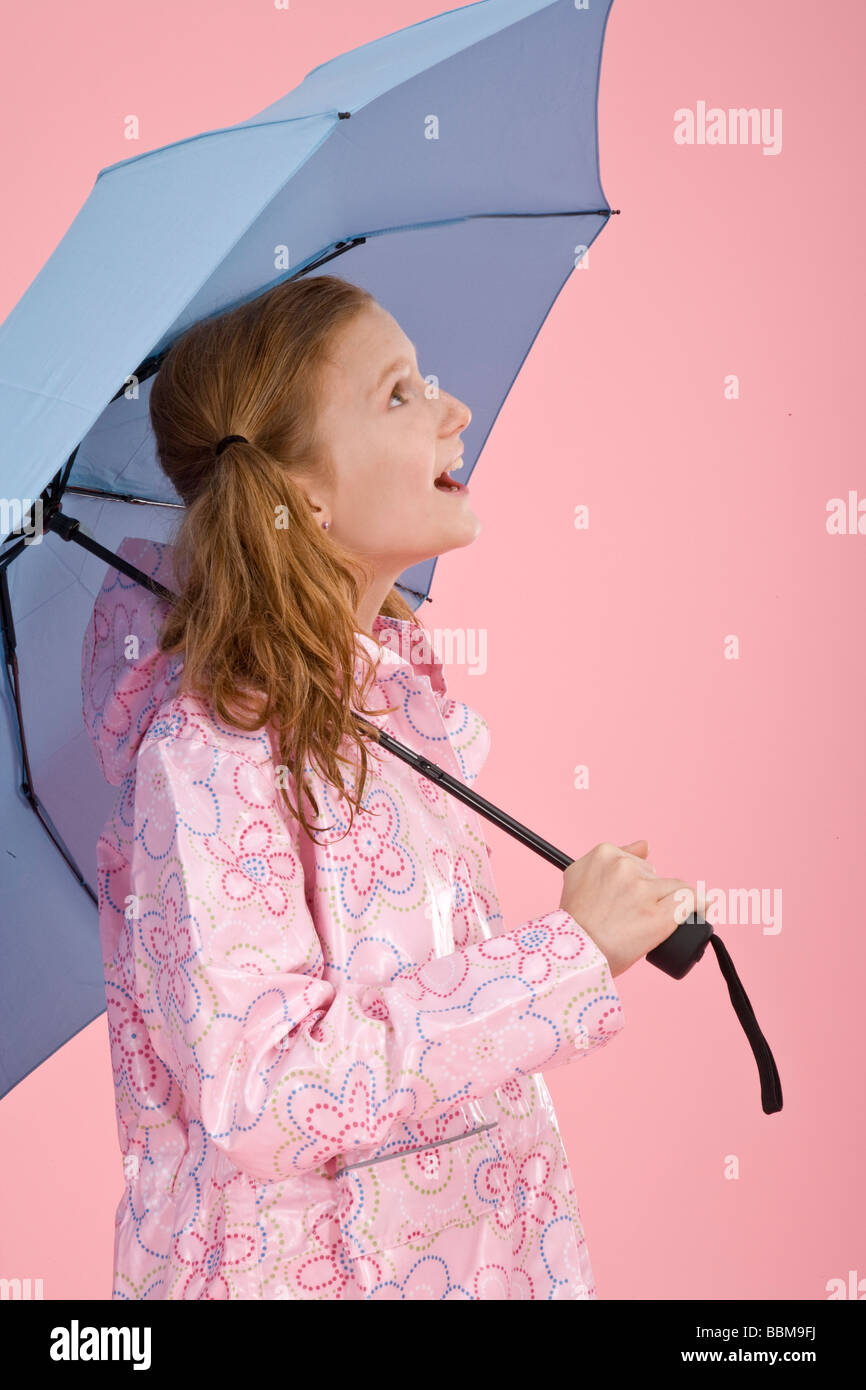 Red-haired girl holding a light blue umbrella and wearing a raincoat in front of a pink backdrop Stock Photo