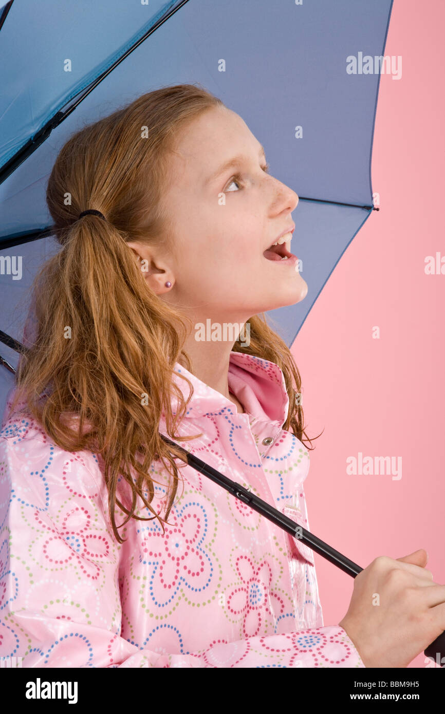 Red-haired girl holding a light blue umbrella and wearing a raincoat in front of a pink backdrop Stock Photo