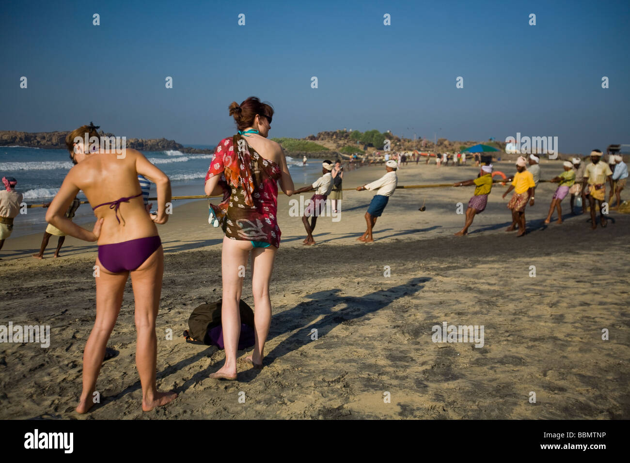 Bikini-clad European women tourists share the beach with traditional local Indian fishermen. Stock Photo