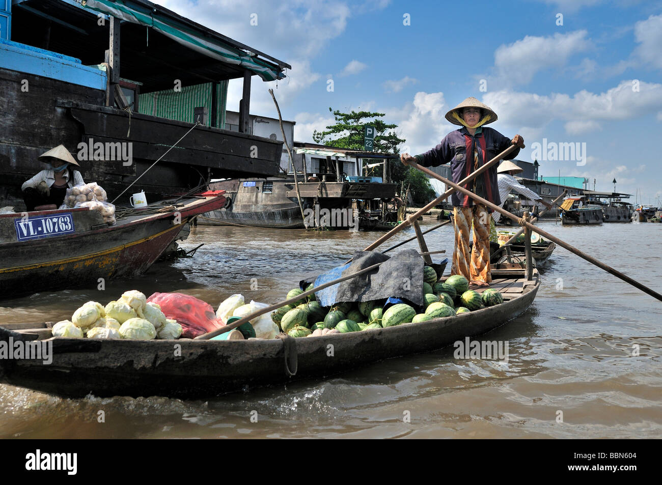 Woman with traditional hat, cone-shaped hat made of palm leaves, standing and rowing a wooden boat on the Mekong, load of fruit Stock Photo