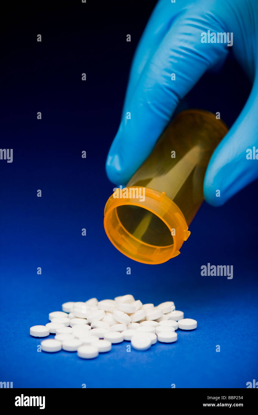 Close-up of a hand in a blue surgical glove holding an empty orange prescription bottle over spilt white pills Stock Photo