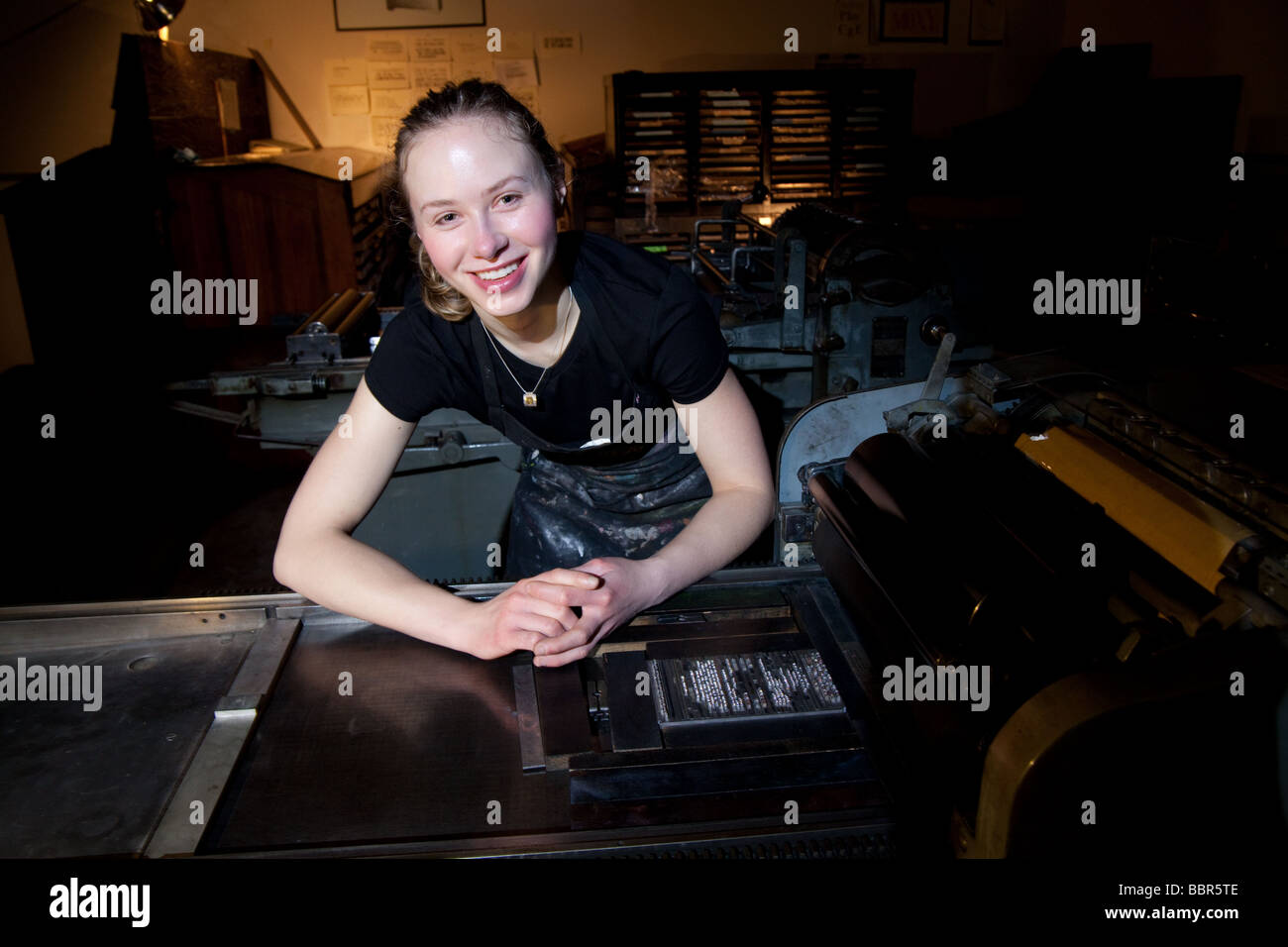 Young white artist woman leaning on a Vandercook press with lead type typeset in a letterpress art studio. Stock Photo