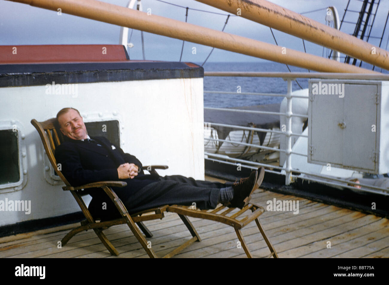 Dressed to travel in style, a dapper business man reclines in a steamer chair at the start of voyage to new life across the pond Stock Photo