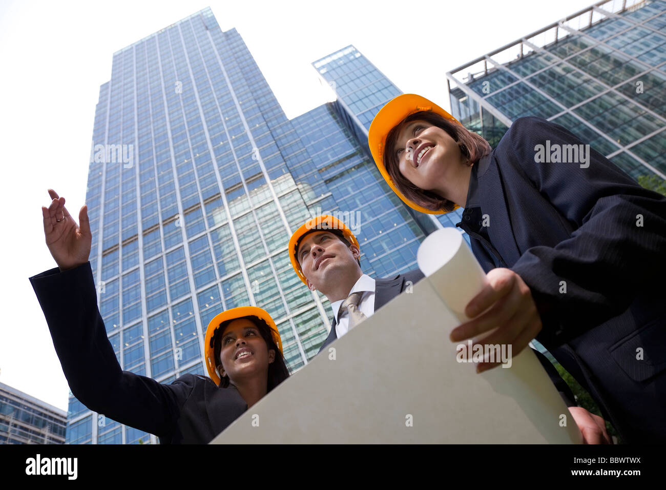 A group of three executives one man and two women wearing hard hats review architectural plans in a modern city environment Stock Photo