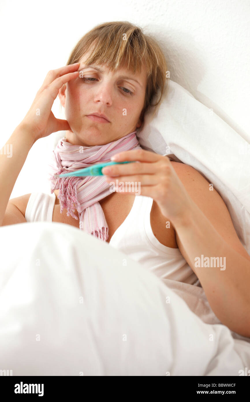 Woman at home, ill with fever in her bed, uses a clinical thermometer for measuring body temperature. Stock Photo