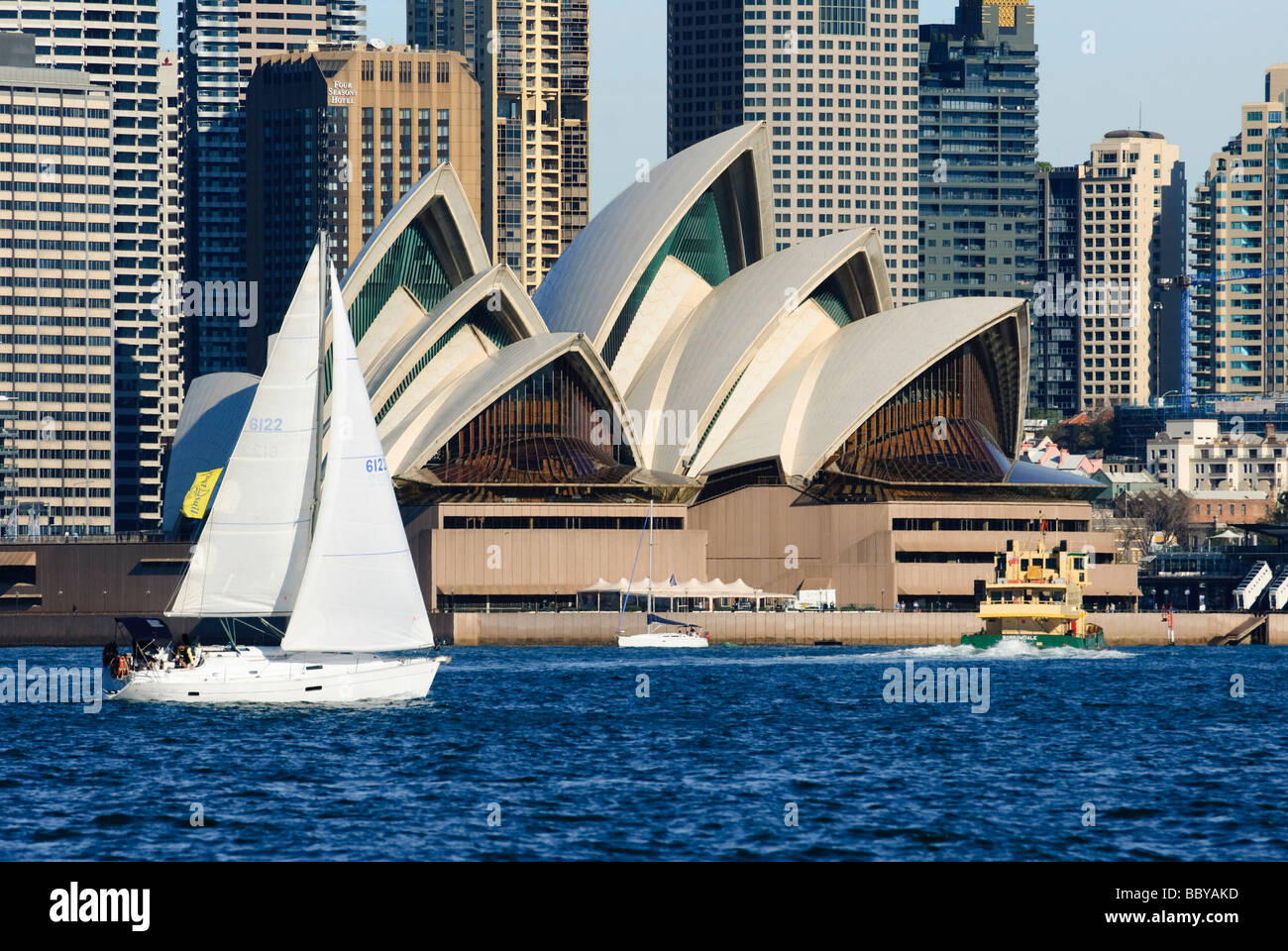 Yacht with white sails and other boats on the blue water of Sydney Harbor, sailing past Sydney Opera House. Stock Photo
