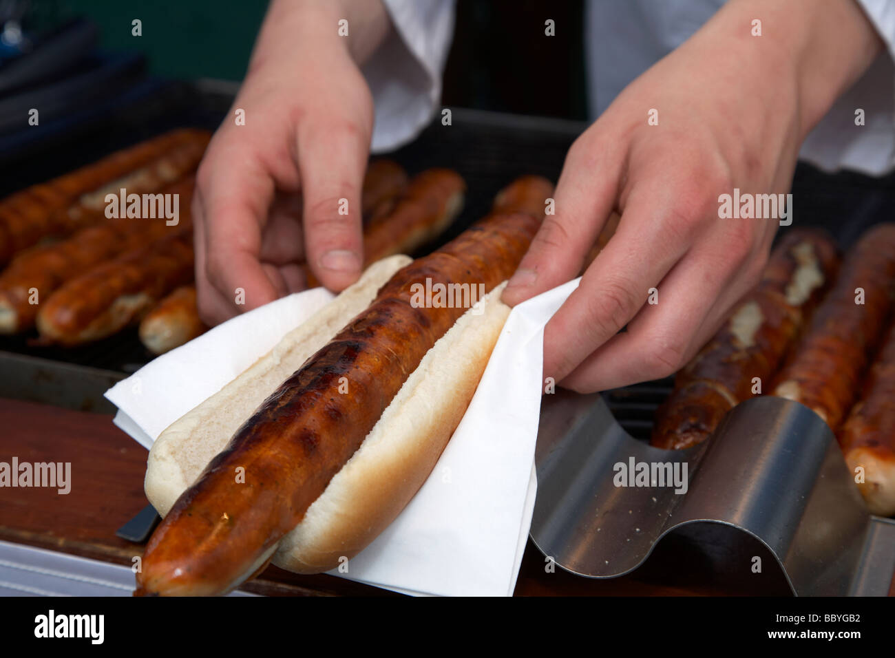 vendor preparing bratwurst in bread roll bap on napkin with german sausages on grill on sale at an outdoor market in the uk Stock Photo
