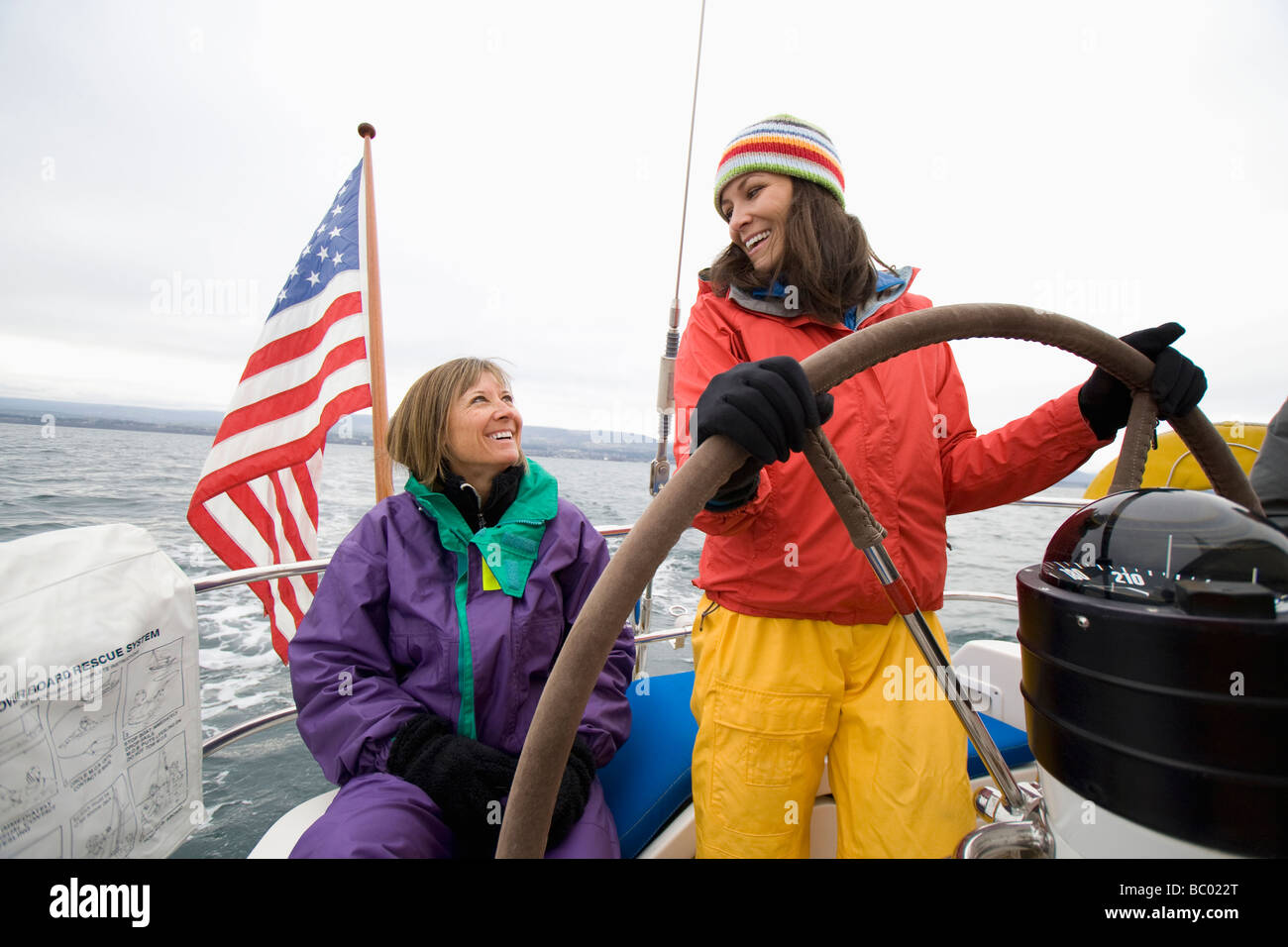 Two smiling women in sailing gear steer a sailboat on a cloudy day