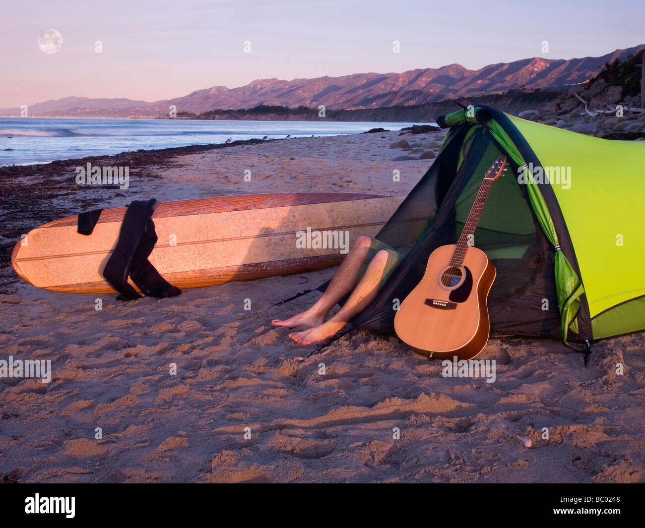 A young man lying in his tent with a guitar while camping on the beach waiting for surf. Stock Photo