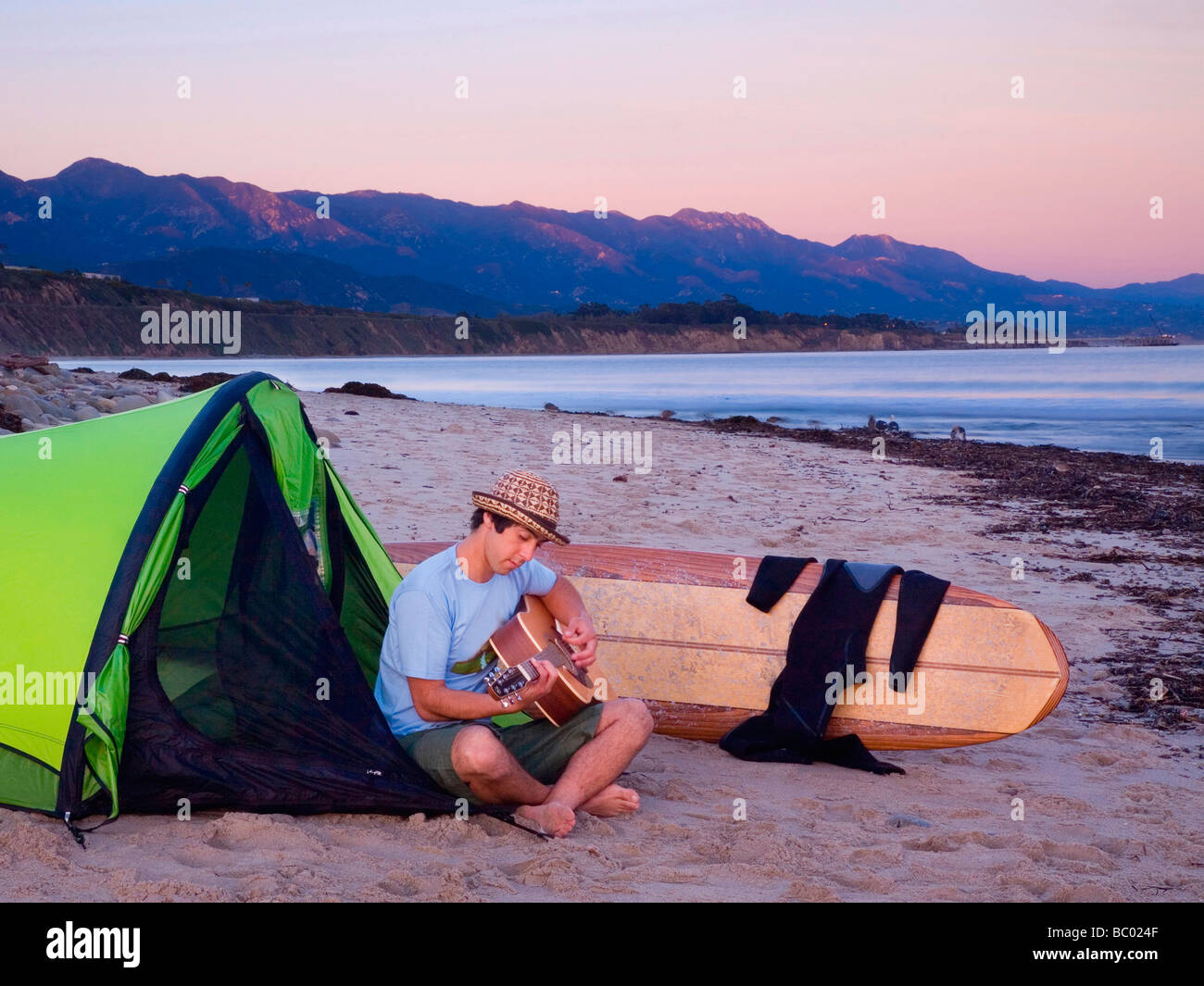 A young man with a guitar sits near his tent while camping on the beach waiting for surf. Stock Photo