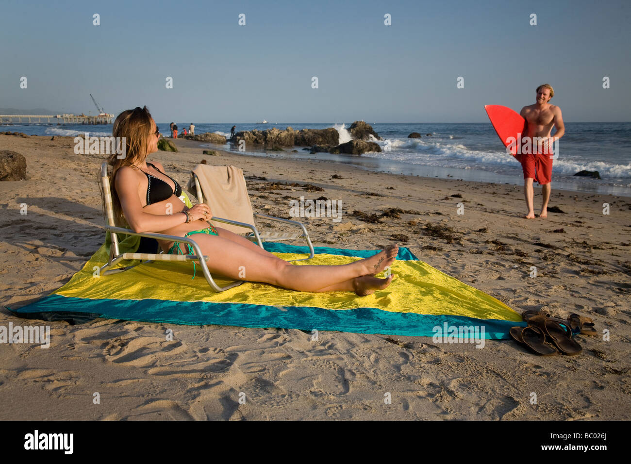 A woman sits in a chair on the beach while a man walks with his surfboard. Stock Photo
