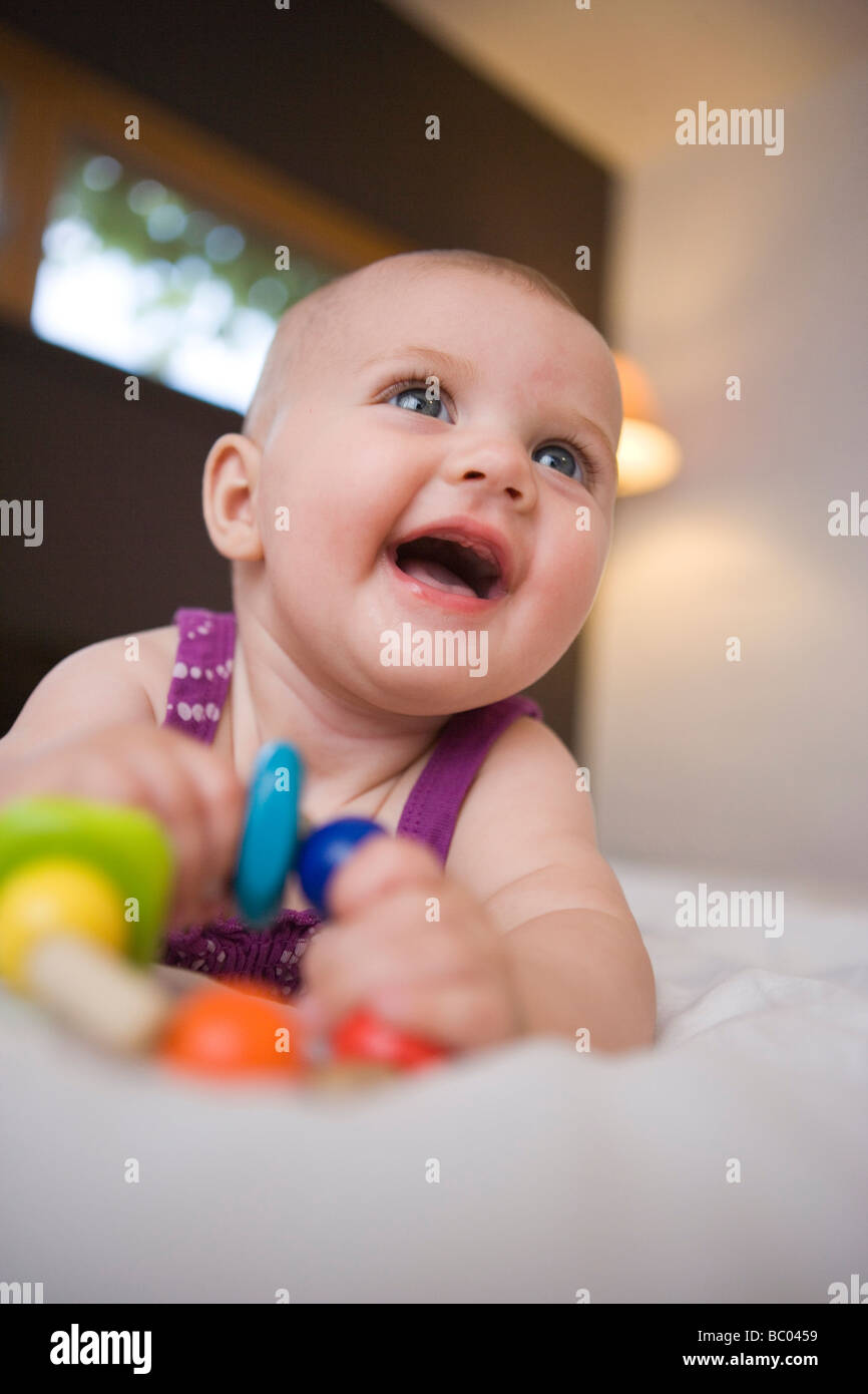 three month old infant girl smiling while holding wooden teething beads, laying on a bed Stock Photo