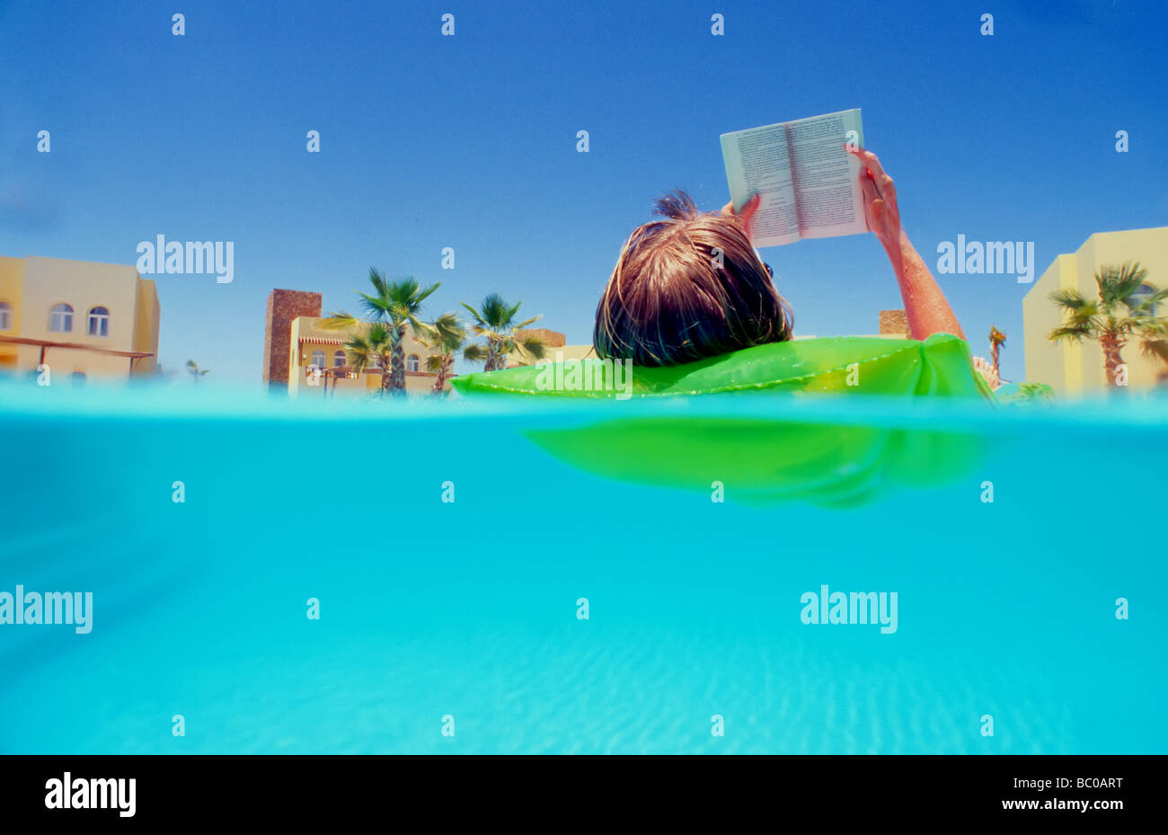 woman 40 floating on inflatable airbed on clear blue water swimming pool reading a book palm trees background Stock Photo