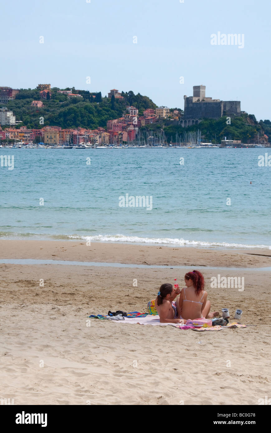 A couple lie on the beach at San Terenzo, Ligurian coast in the gulf of Poets near La Spezia with Lerici in the distance Stock Photo