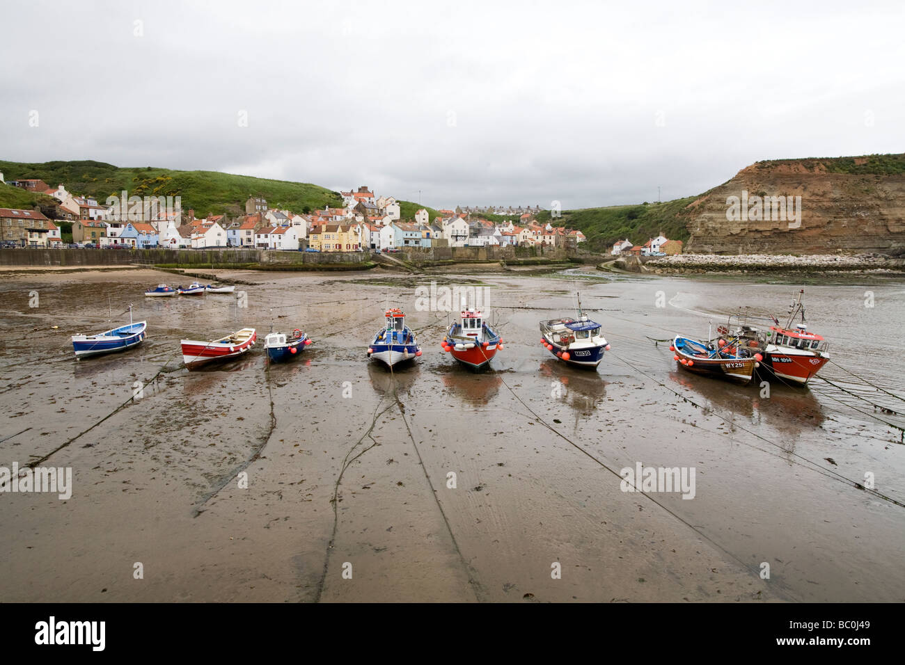 Small tied-up boats in a row left dry at low tide standing on sand with reflections Staithes North Yorkshire England Stock Photo