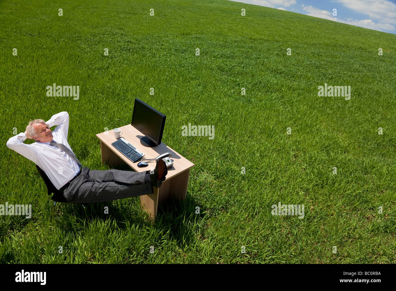 Business concept shot showing an older male executive relaxing at his desk in a green field with a blue sky Stock Photo