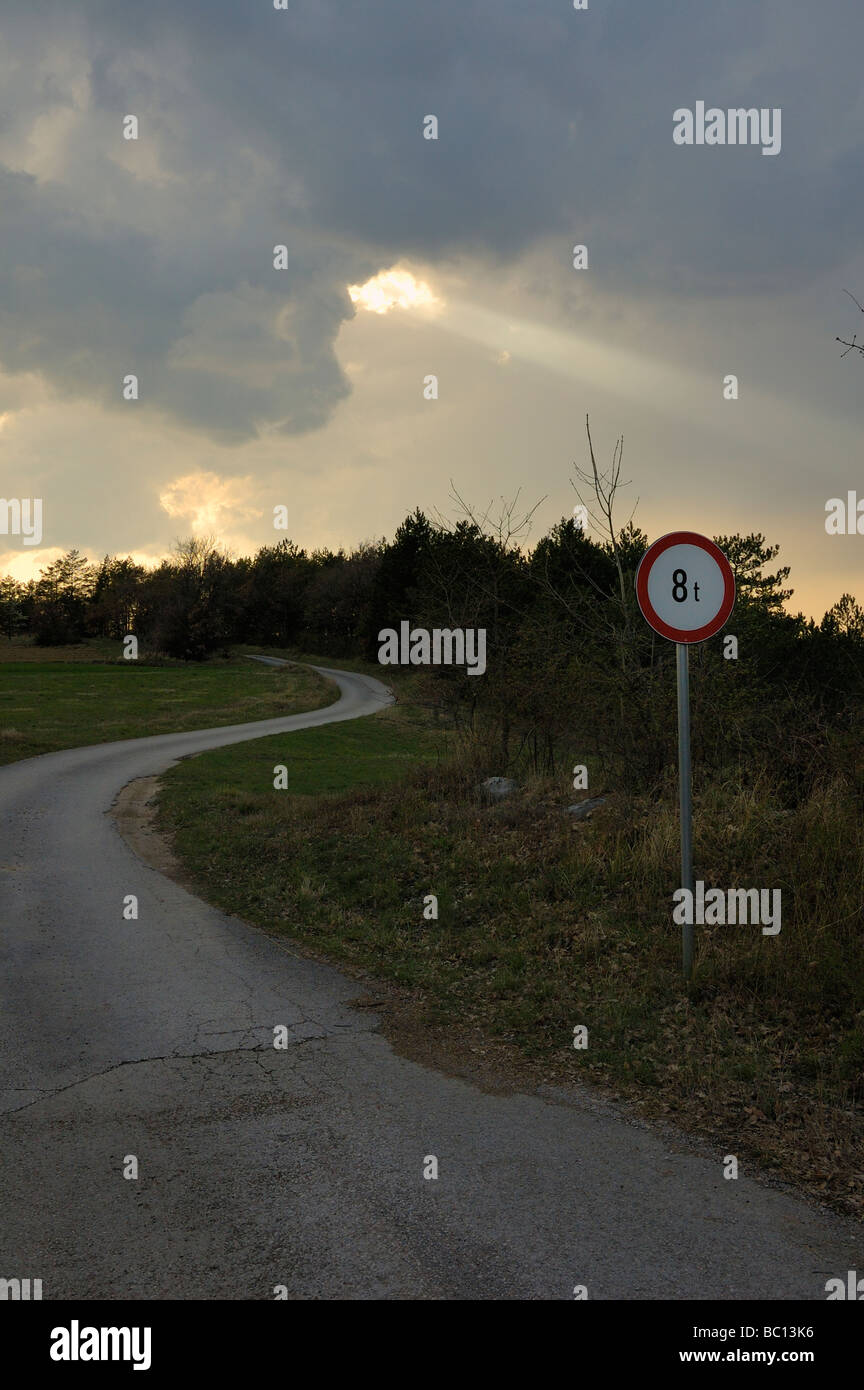 Country road curve with weight limit traffic sign and sun ray through the clouds in background Stock Photo