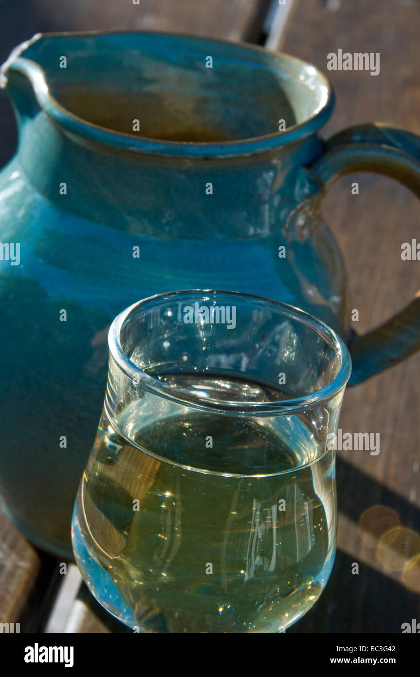 Sunlit glass of simple country white wine on terrace table with blue earthenware jug carafe behind Stock Photo