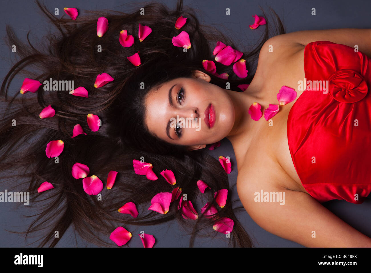 Beautiful brunette woman in a red dress lying down with pink rose petals in her hair Stock Photo