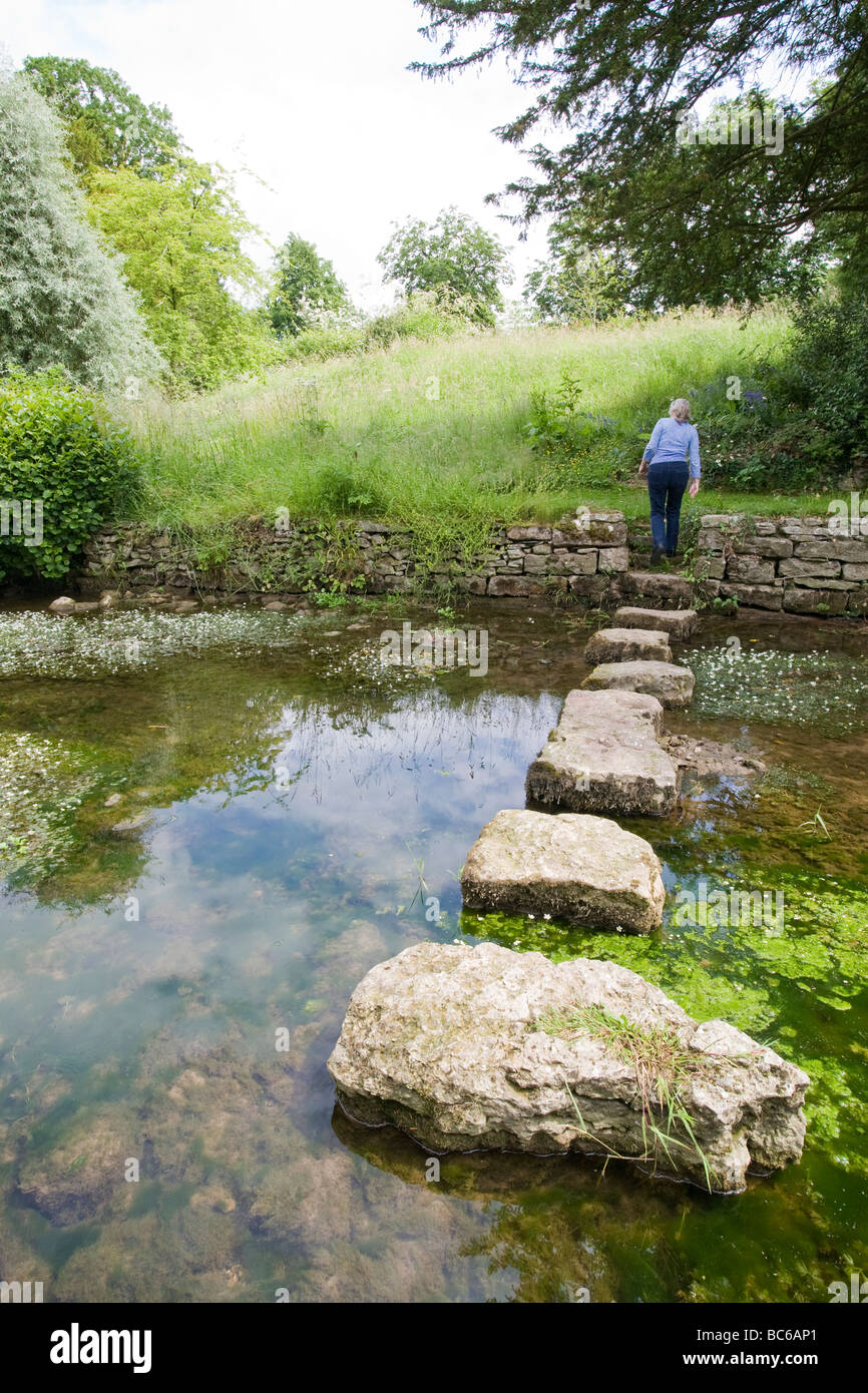 Back view of female at the end of a series of stepping stones over a small stream with grass and tress in distance Stock Photo