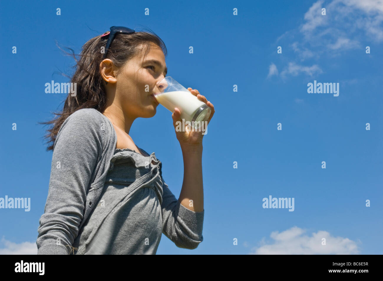 A young caucasian girl (12) drinking a glass of fresh milk on a sunny day with a blue sky background. Stock Photo