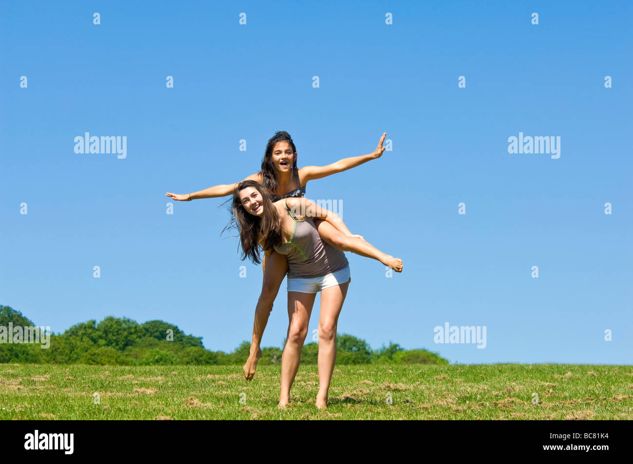 Horizontal portrait of two teenage girls having fun having a piggy back ride in a park on a bright sunny day Stock Photo
