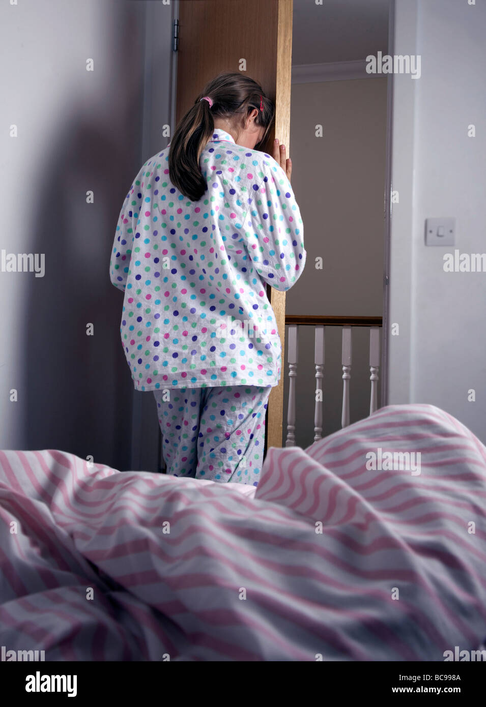 A young pony tailed girl wearing pyjamas, buries her head behind the bedroom door. Stock Photo