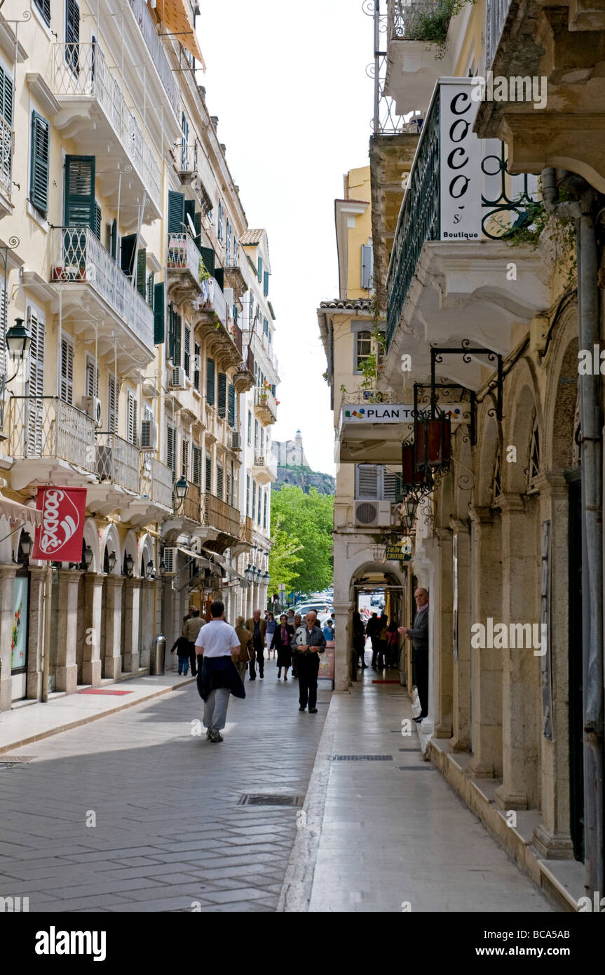 Tall old buildings with green louvered window shutters and balconies enclose a narrow cobblestone street in Corfu Town Stock Photo