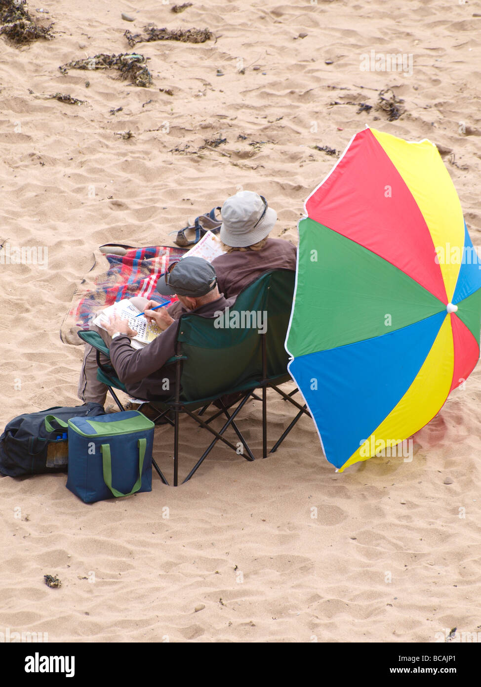 Old couple at the beach with large sun umbrella doing crosswords Stock