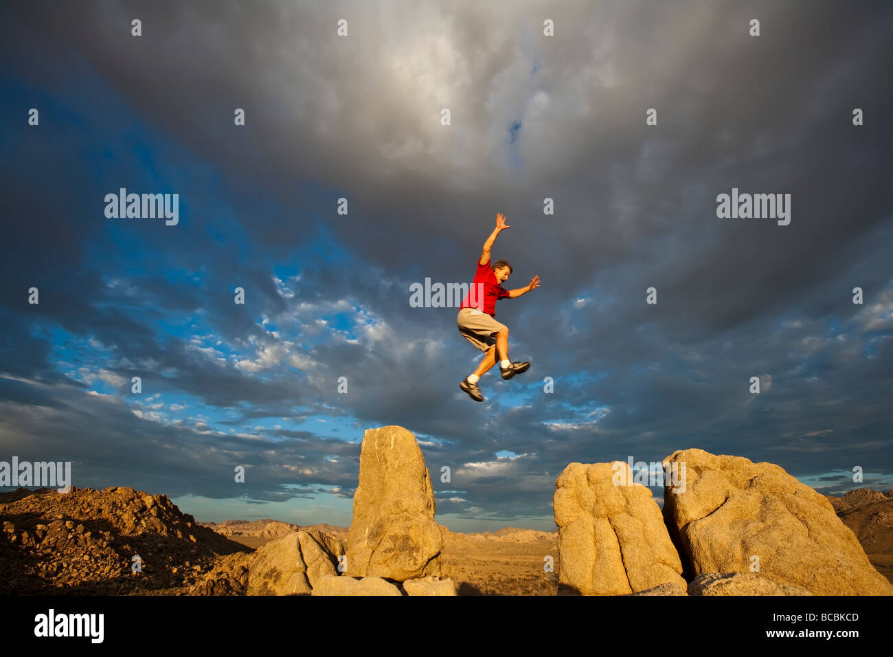 Rock climber leaps across a gap on the summit of a pinnacle after a successful ascent as storm clouds build in the setting sun Stock Photo