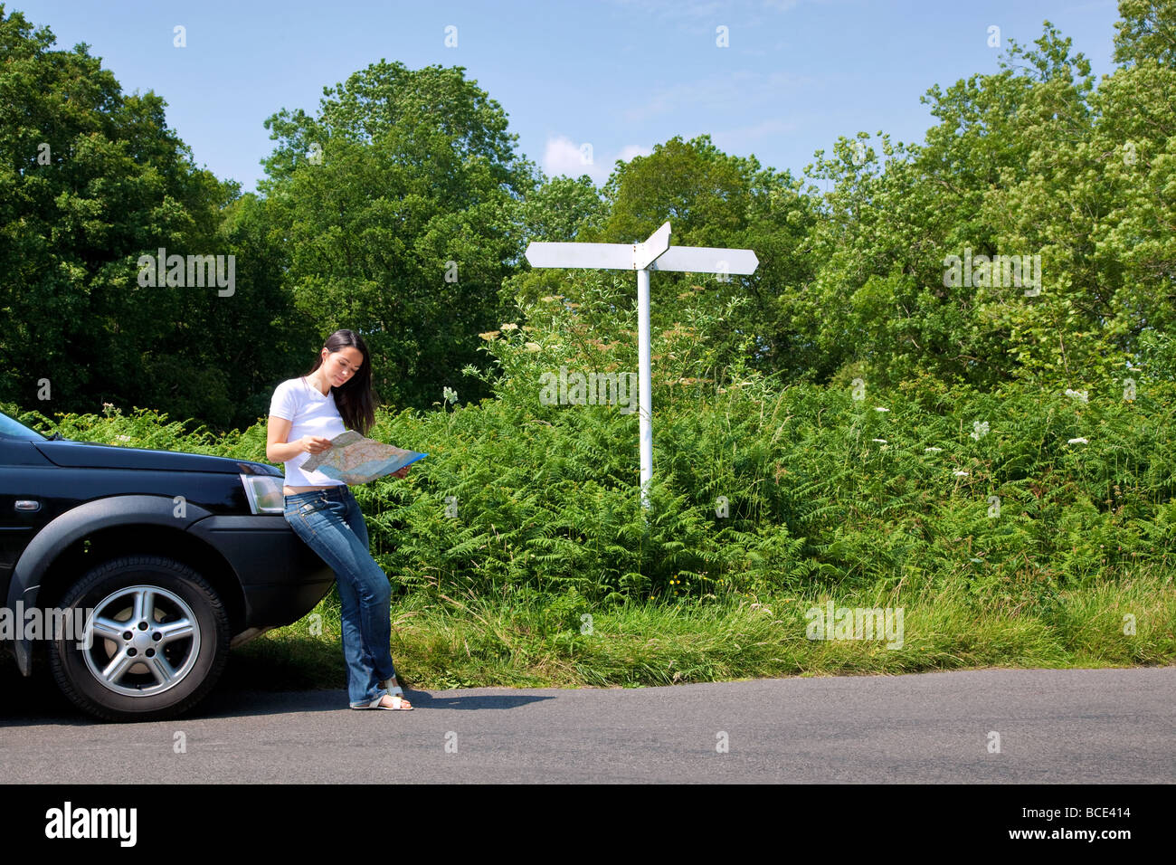 A woman leaning on her car as she reads a map next to a roadside sign the sign is blank for you to add your own text Stock Photo