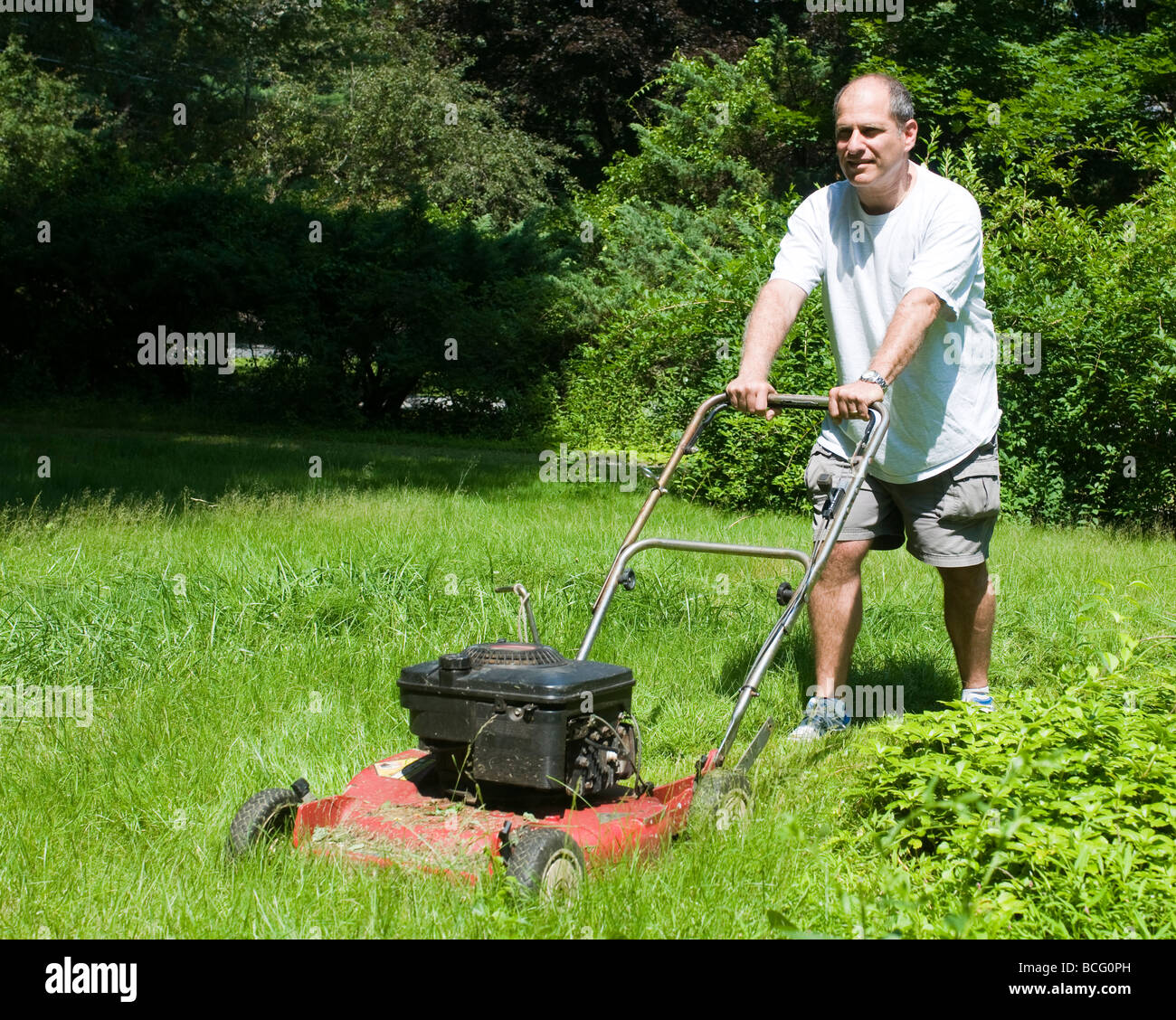 handsome middle age man cutting overgrown grass with old fashioned lawn mower at suburban house Stock Photo