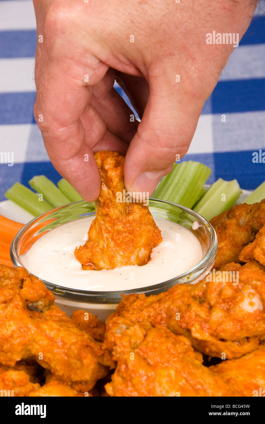A dish of chicken hot wings celery and carrots with dipping sauce attracts a man who dips a wing into some tasty ranch sauce Stock Photo