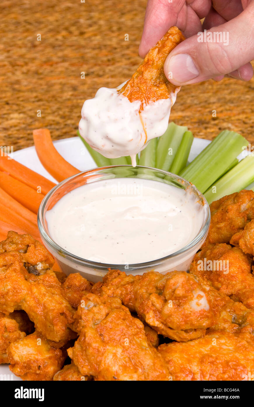 A dish of chicken hot wings celery and carrots with dipping sauce attracts a man who dips a wing