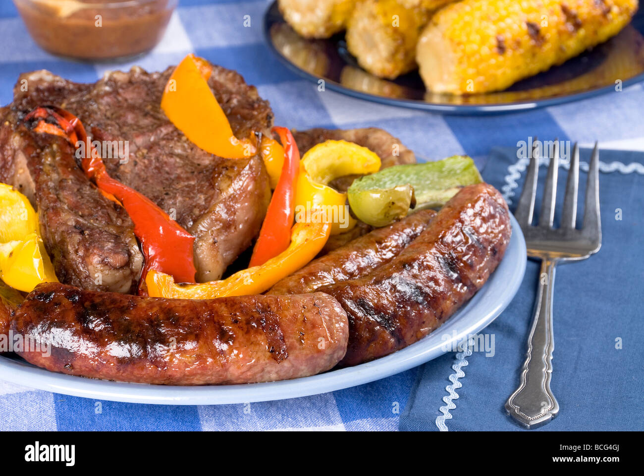 A bratwurst and steak dinner with corn on the cob on a picnic table and tablecloth Stock Photo