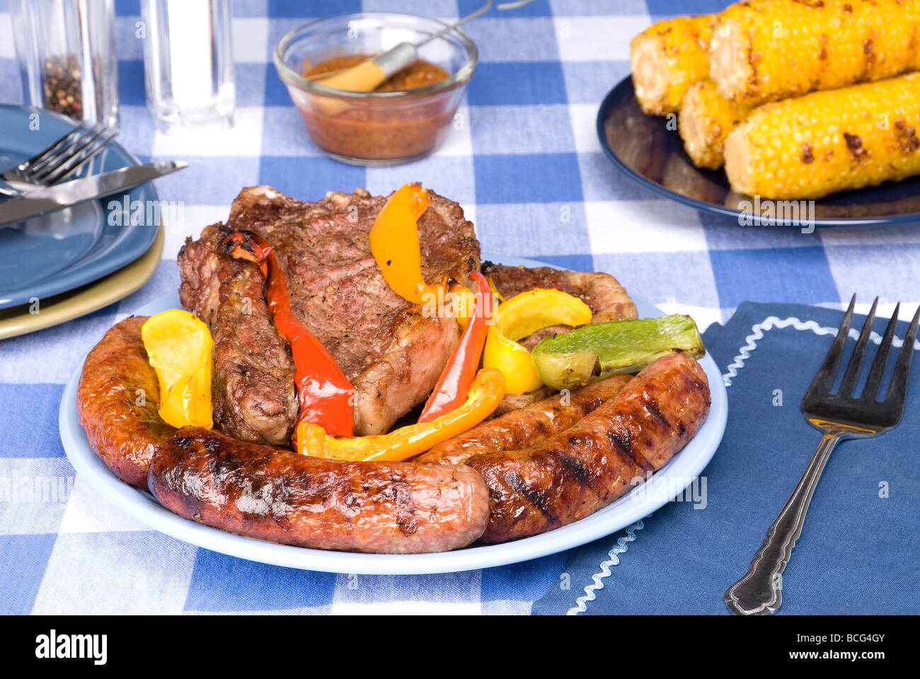 A bratwurst and steak dinner with corn on the cob on a picnic table and tablecloth Stock Photo