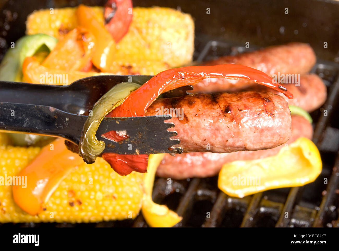 A hot beer bratwurst with cooked bell peppers against a barbecue gripp of corn on the cob and other meat Stock Photo