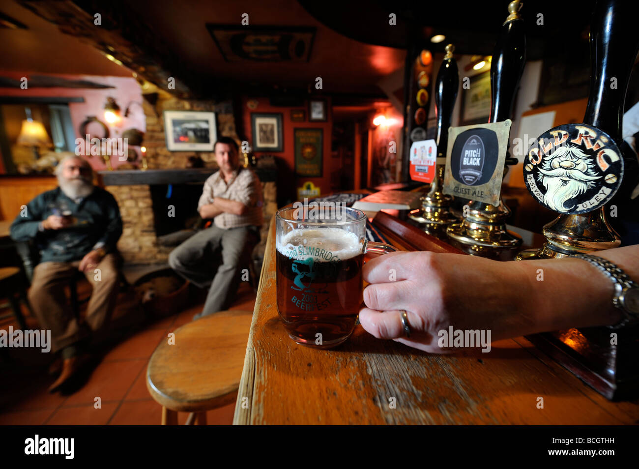TWO MEN SIT BY AN OPEN FIRE IN A TRADITIONAL PUB AS THE LANDLADY PLACES A PINT OF ALE ON THE BAR GLOUCESTERSHIRE ENGLAND UK Stock Photo