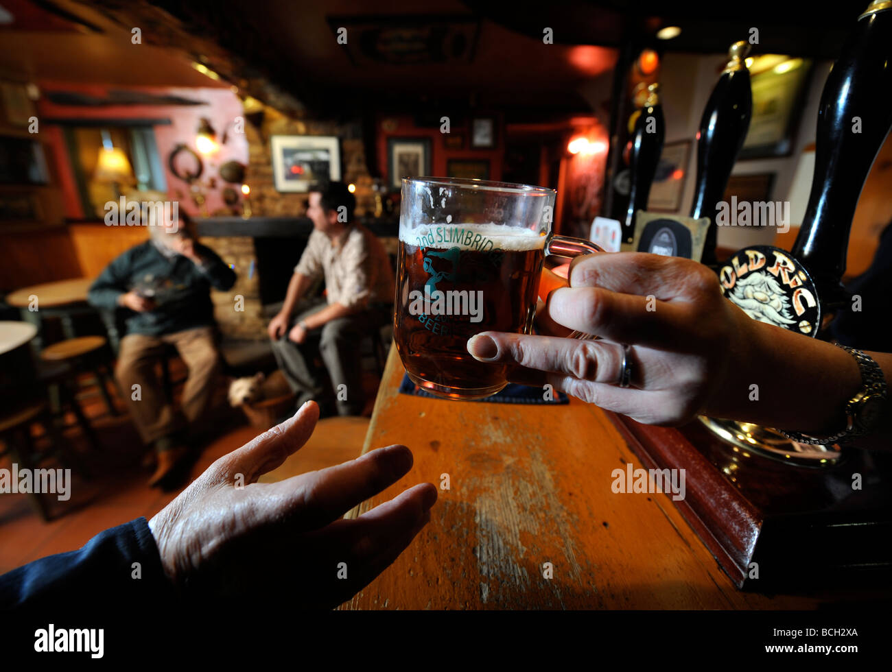 TWO MEN SIT BY AN OPEN FIRE IN A TRADITIONAL PUB AS THE LANDLADY SERVES A PINT OF ALE OVER THE BAR GLOUCESTERSHIRE ENGLAND UK Stock Photo
