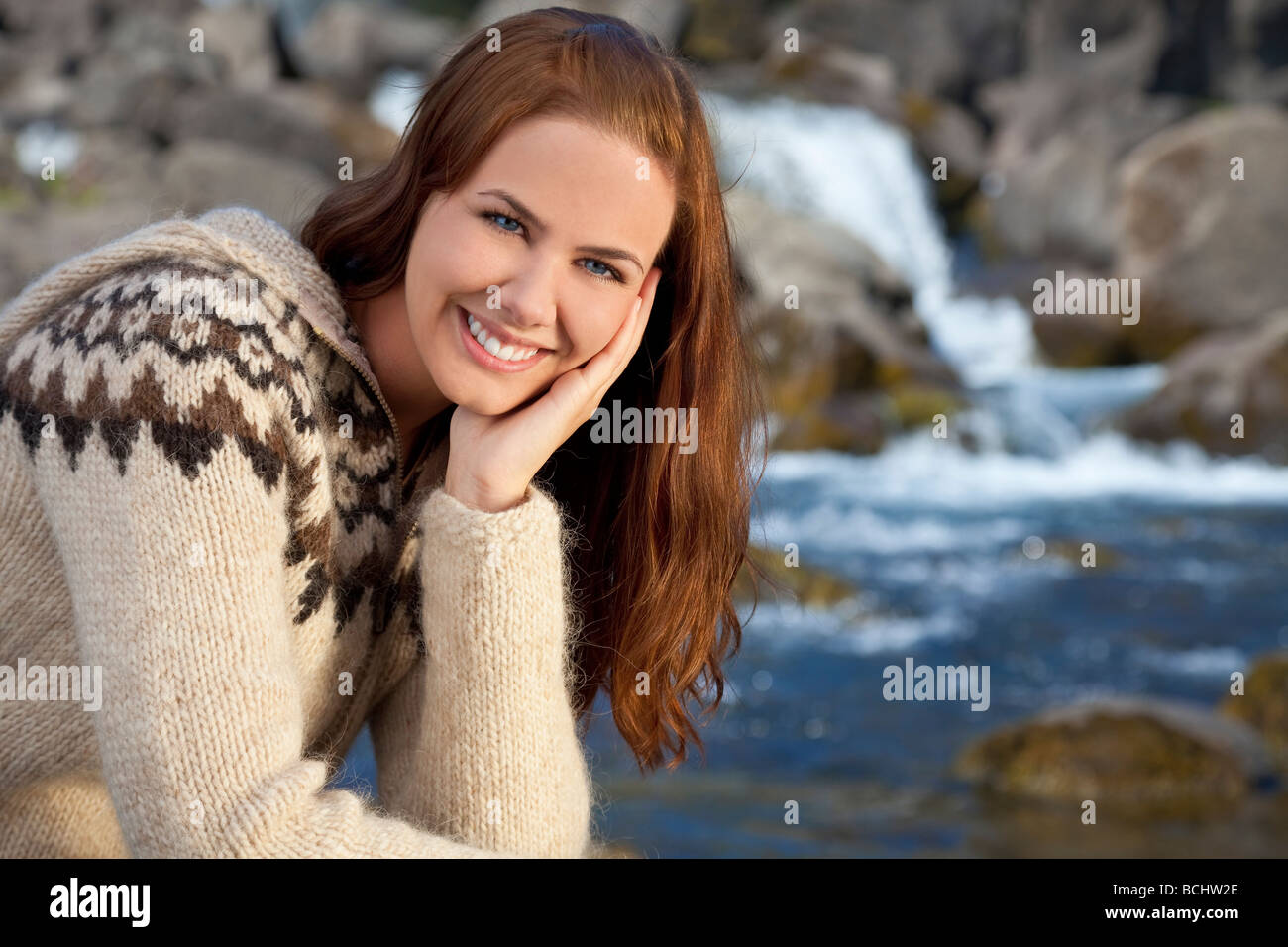 A beautiful Scandinavian woman wearing traditionally patterned knitwear sitting and relaxing by a flowing mountain stream Stock Photo