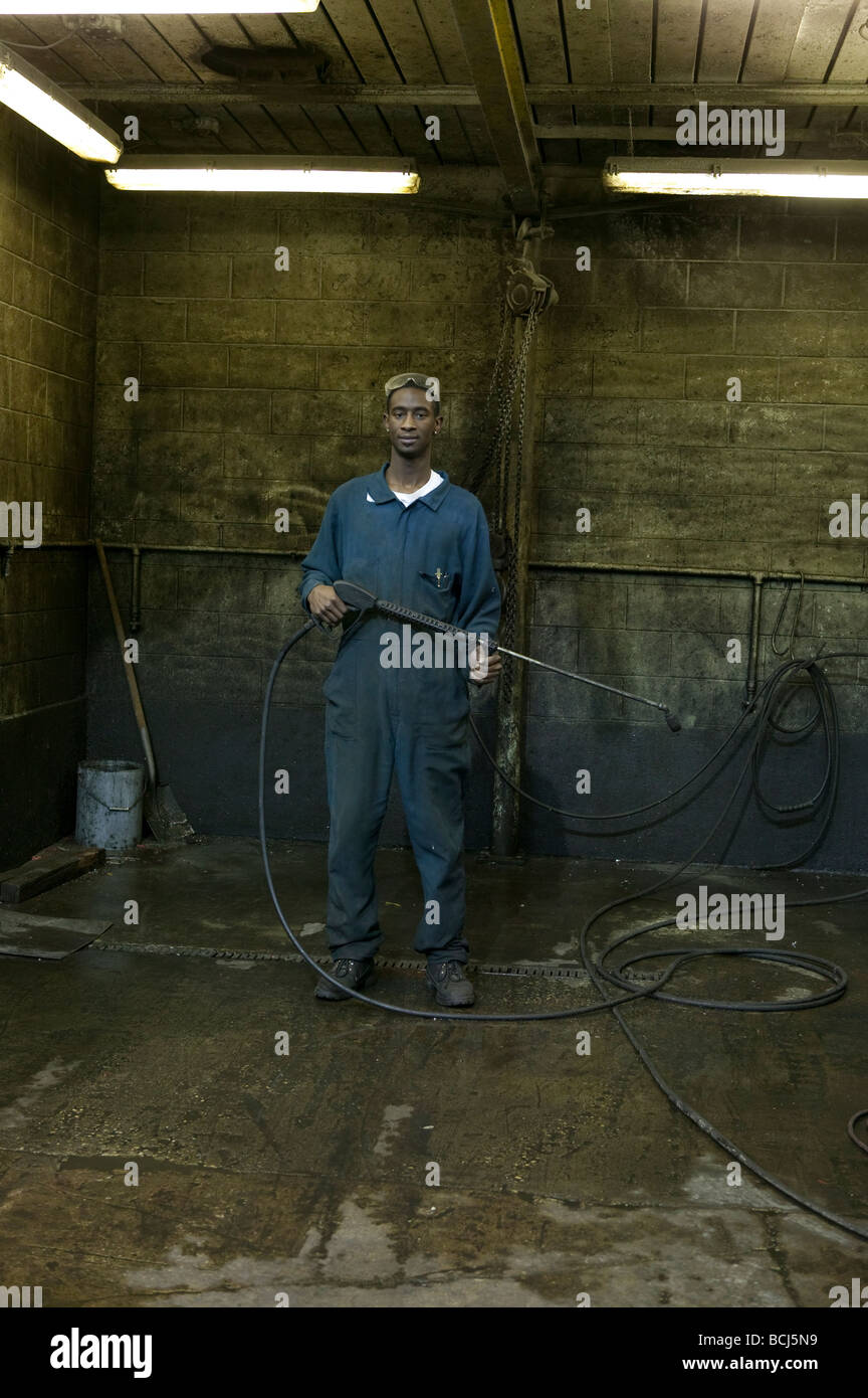 African American male in blue coveralls overalls holding steam cleaning wand device standing in dirty work bay Stock Photo