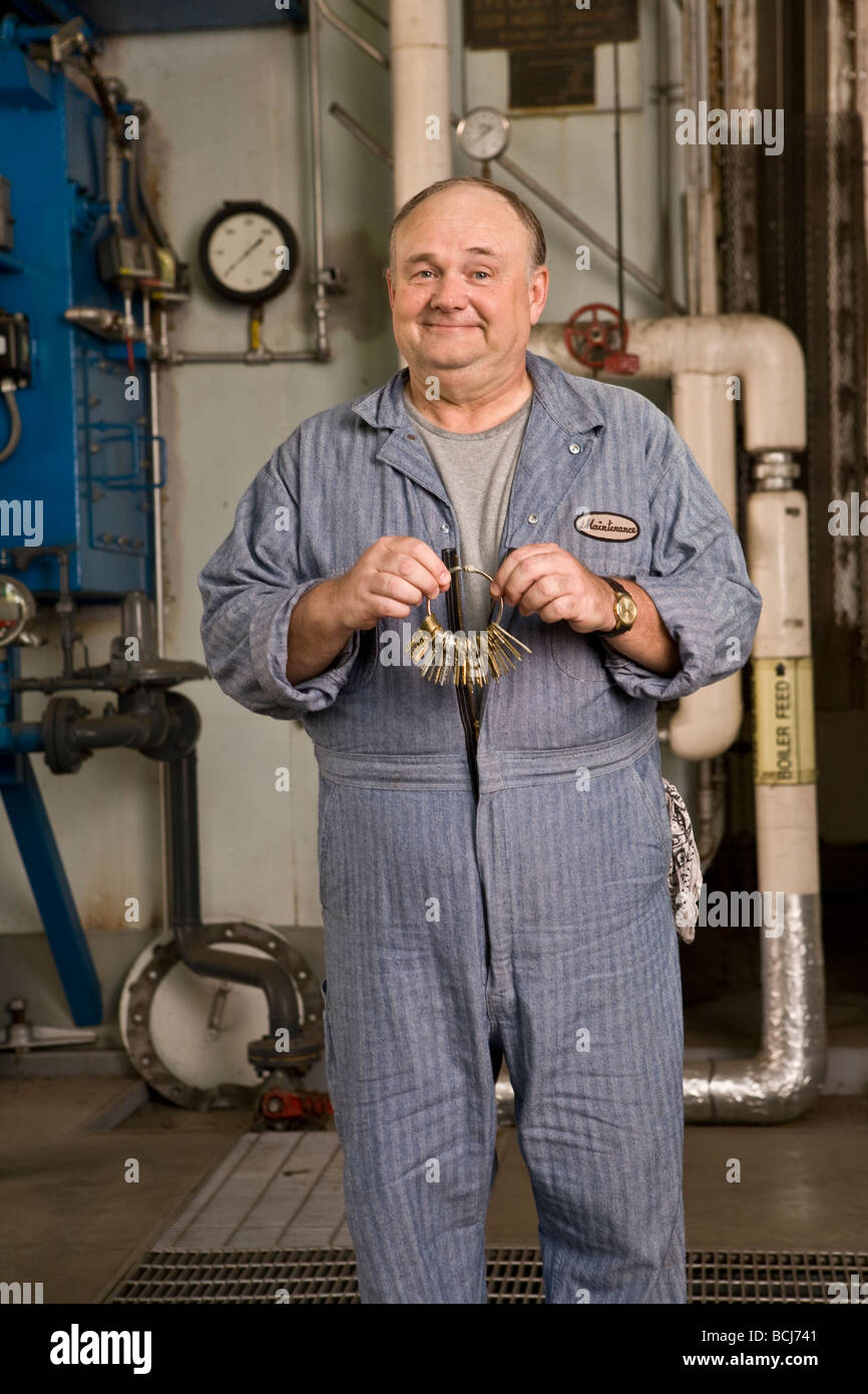 Male Caucasian in blue custodian/janitor overalls in factory setting including boiler, pipes, valves.  He is holding set of keys Stock Photo