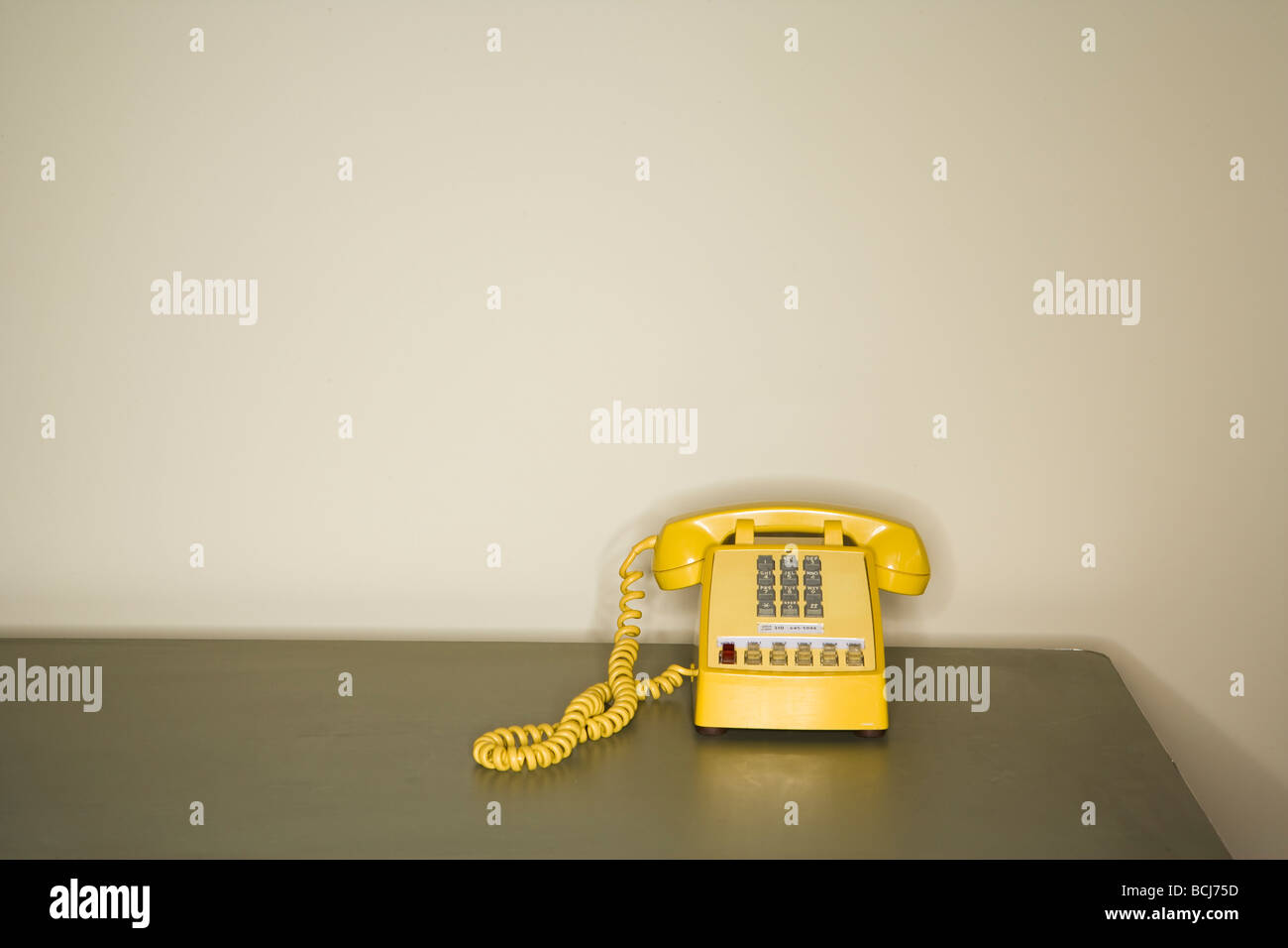 Yellow touch tone telephone sitting on metal desk in front of white wall. Stock Photo