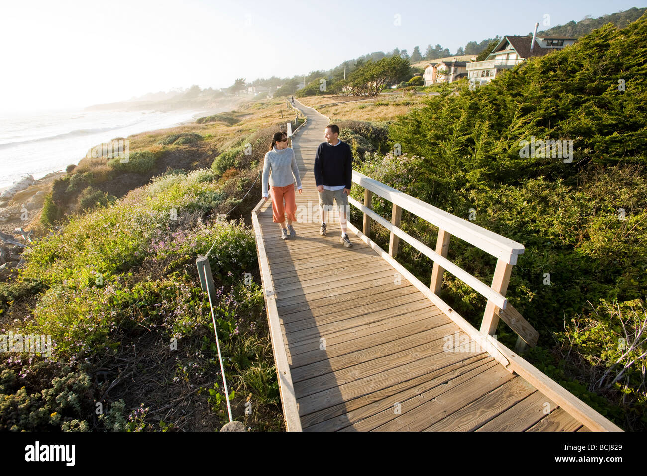 Male and female couple walking on wooden boardwalk path along cliffs beach in Cambria California with view of Pacific Ocean Stock Photo