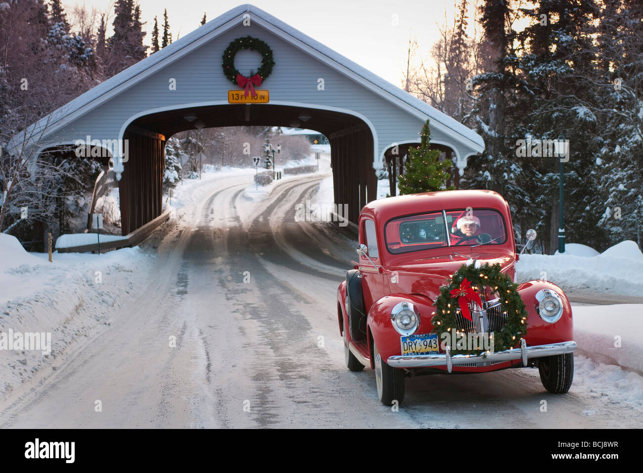 Man driving a vintage Ford pickup through a covered bridge with a Christmas wreath on the grill and a tree in the back, Alaska Stock Photo