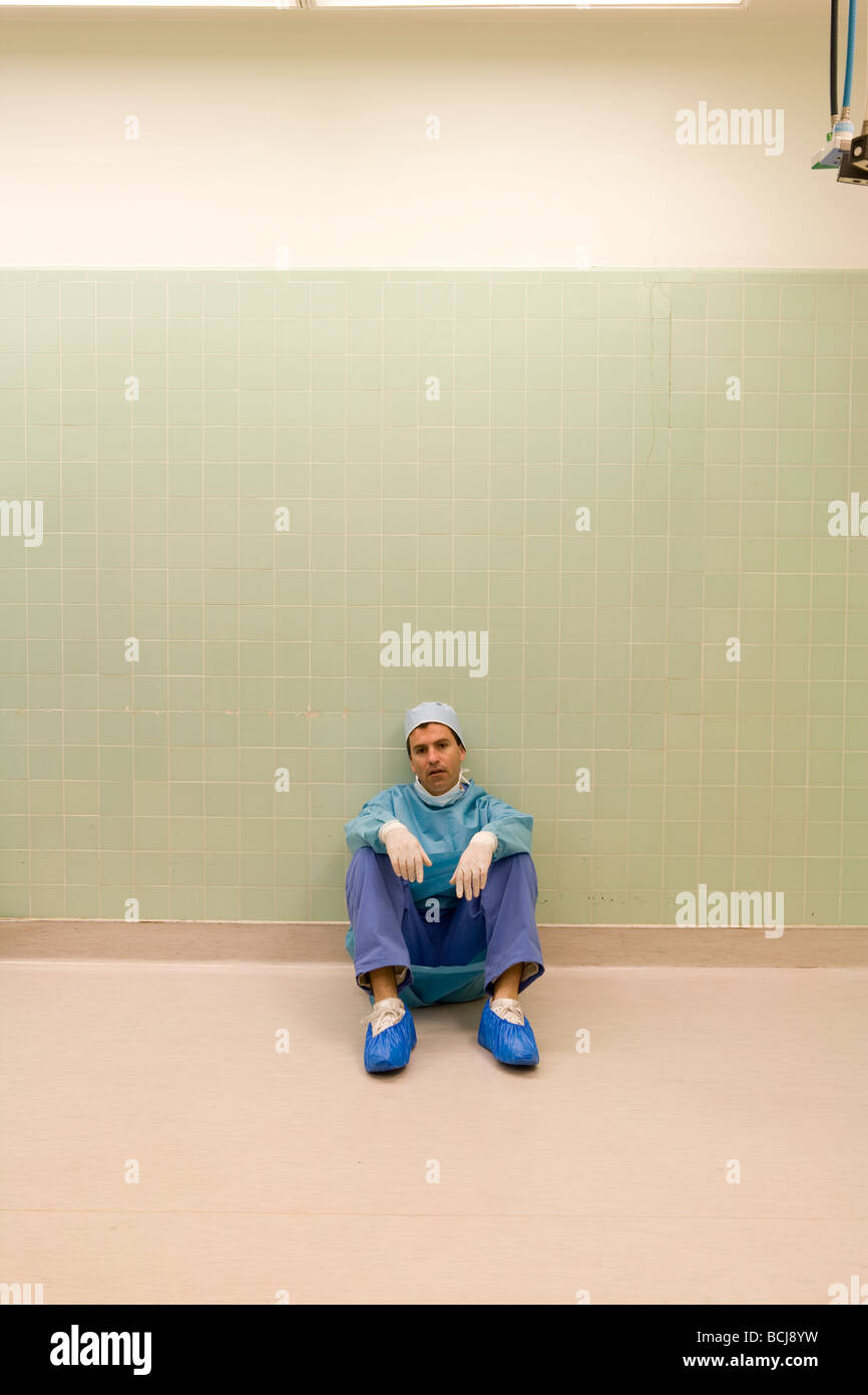 Caucasian male surgeon/doctor in blue surgical scrubs sitting on floor against green tile wall, tired, exhausted. Stock Photo