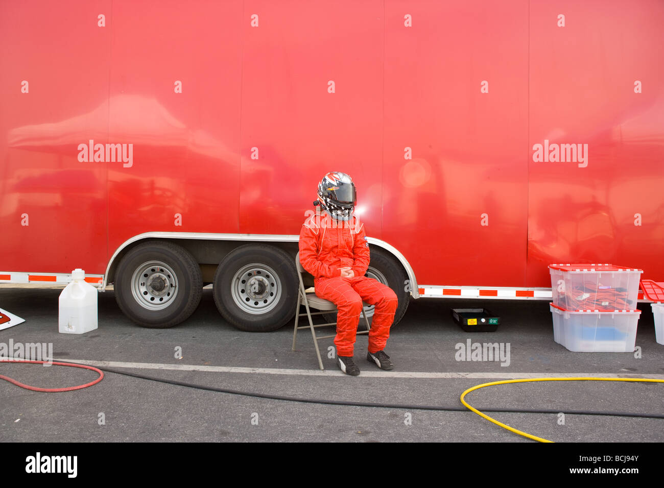 Race car driver wearing helmet and red jump suit sitting in chair in front of red transport vehicle  Stock Photo