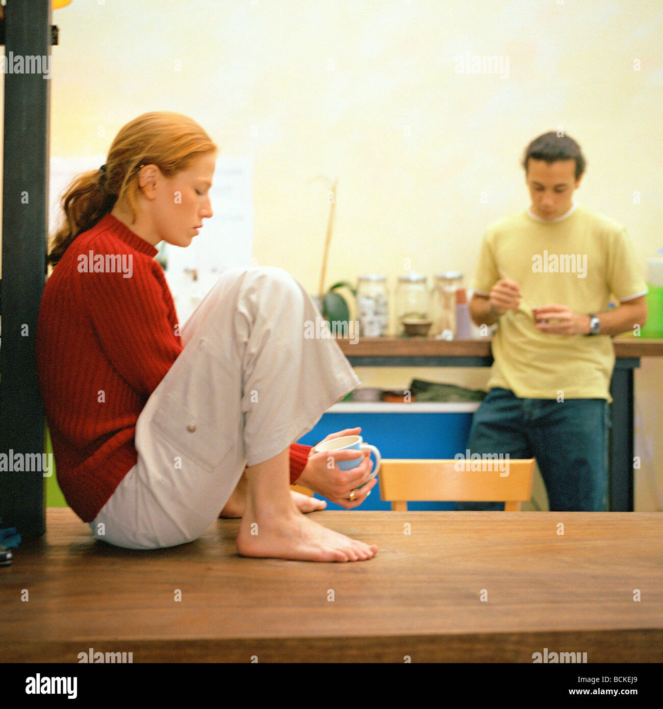 Young woman sitting on kitchen table, man standing with cup Stock Photo